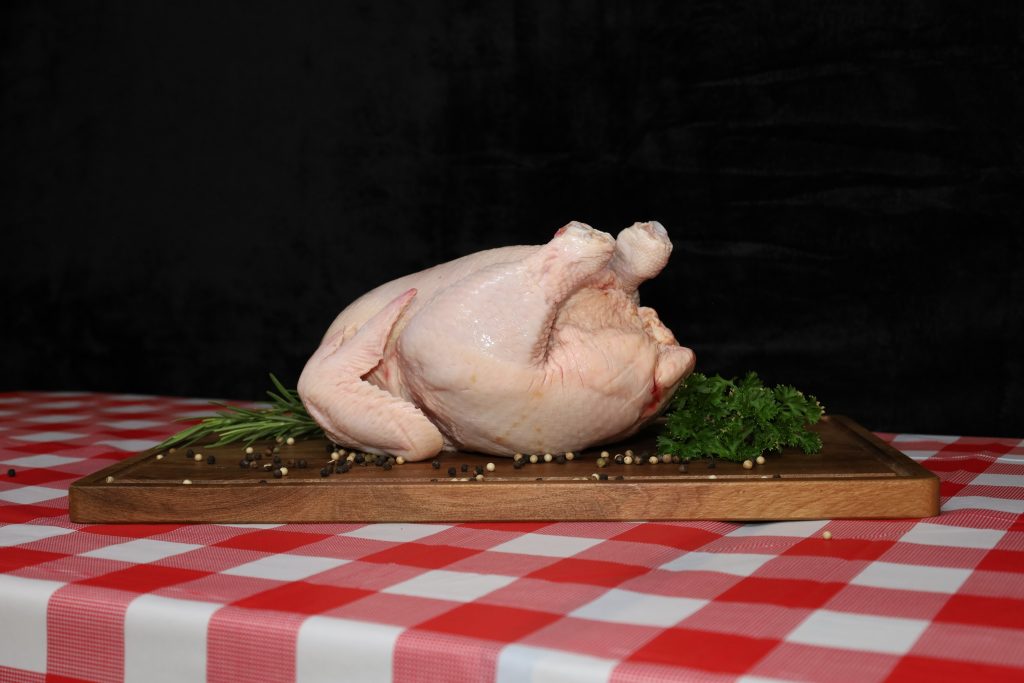 Raw whole free-range chicken on wooden board with fresh herbs on red and white checkered tablecloth.